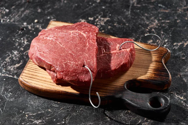 Planche à découper en bois avec steaks de boeuf crus sur marbre noir — Photo de stock