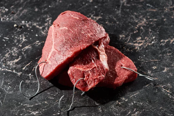 Raw beef steaks with metal hooks on black marble surface — Stock Photo