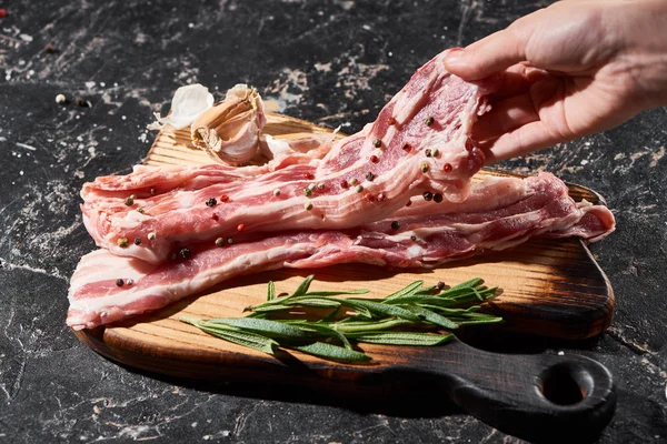 Cropped view of raw pork slice in hand near rosemary and garlic on black marble surface — Stock Photo