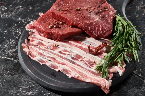 Wooden pan with raw beef steaks and bacon slices near rosemary and dried cayenne pepper on black marble surface — Stock Photo