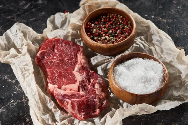 Parchment paper with raw meat fillet near wooden bowls with salt and pepper on black marble surface — Stock Photo