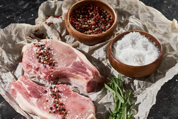 Raw pork steaks on parchment paper near wooden bowls with salt and pepper on black marble surface — Stock Photo
