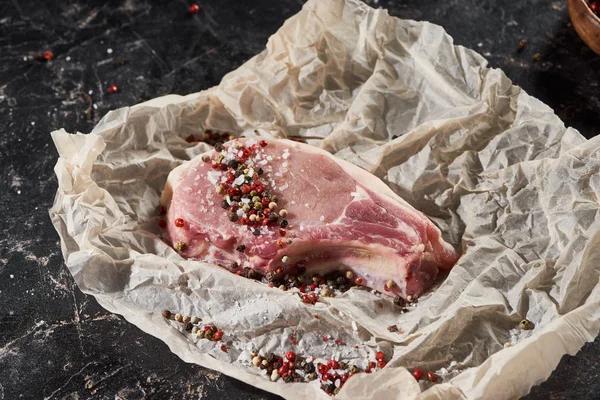 Rohes Schweinesteak mit Salz und Pfeffer auf Pergamentpapier auf schwarzer Marmoroberfläche bestreut — Stockfoto