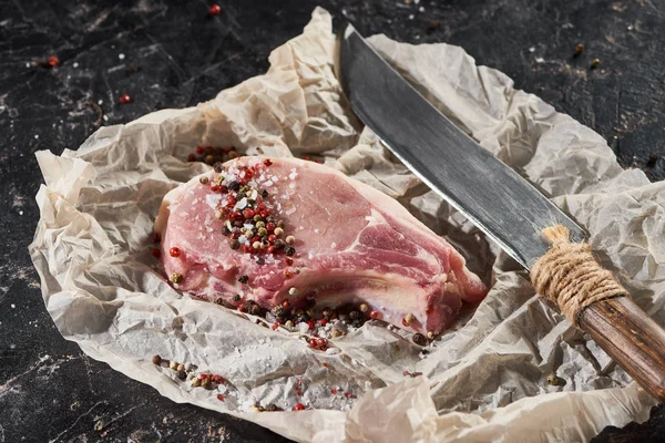 Steak de porc cru saupoudré de sel et de poivre près du couteau sur du papier sulfurisé sur une surface de marbre noir — Photo de stock