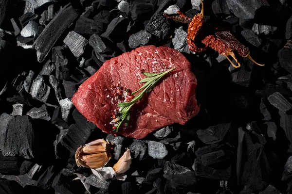 Vue du dessus du steak de boeuf cru au romarin, sel et poivre près des gousses d'ail et de la Cayenne séchée sur les charbons — Photo de stock