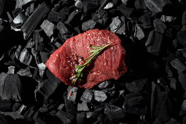 Top view of raw beef steak with rosemary, salt and pepper on charcoals — Stock Photo