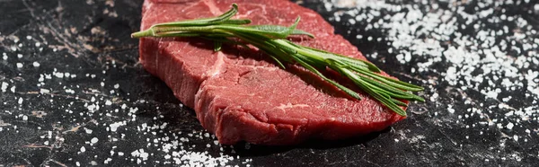 Panoramic shot of unprocessed beef steak with rosemary on black marble surface with scattered salt — Stock Photo