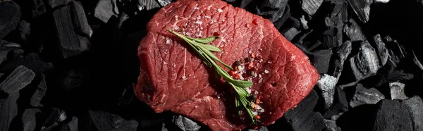 Panoramic shot of raw beef sirloin with rosemary, salt and pepper on coal pieces — Stock Photo