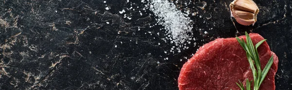 Panoramic shot of raw beef steak with rosemary twig near garlic and scattered salt on black marble surface — Stock Photo