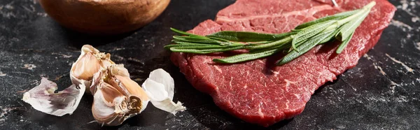 Panoramic shot of uncooked beef sirloin with rosemary sprig near garlic on black marble surface — Stock Photo