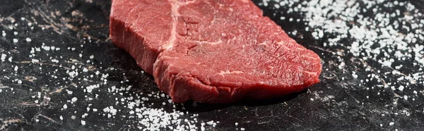 Panoramic shot of fresh raw beef steak on black marble surface with scattered salt — Stock Photo
