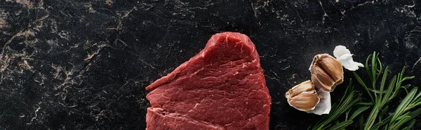 Панорамний знімок зубчиків часнику, гілок розмарину та сирого яловичого стейка на поверхні чорного мармуру — стокове фото