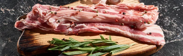 Plano panorámico de tabla de cortar de madera con rodajas de cerdo en bruto espolvoreado con granos de pimienta sobre la superficie de mármol negro - foto de stock