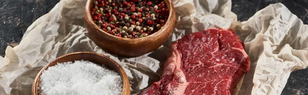 Panoramic shot of raw beef fillet near wooden bowls with salt and peppercorns on parchment paper — Stock Photo