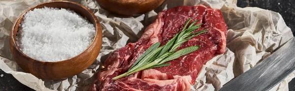 Panoramic shot of raw beef fillet with rosemary twig near wooden bowl with salt on parchment paper — Stock Photo