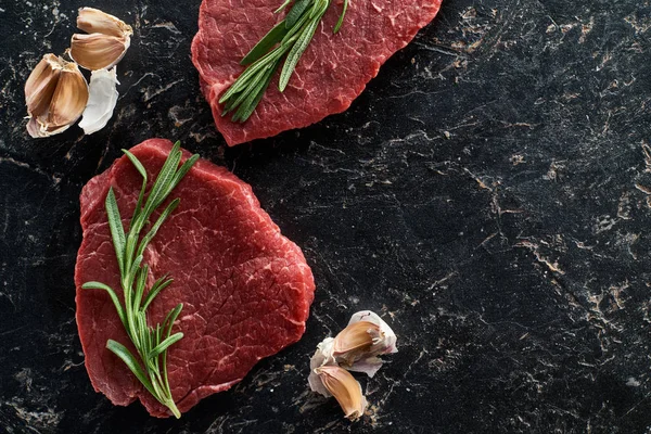 Top view of uncooked beef sirloins with garlic and rosemary on black marble background — Stock Photo