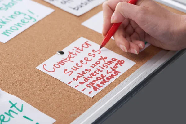 Cropped view of woman writing competitor success notes on card pinned on cork board — Stock Photo