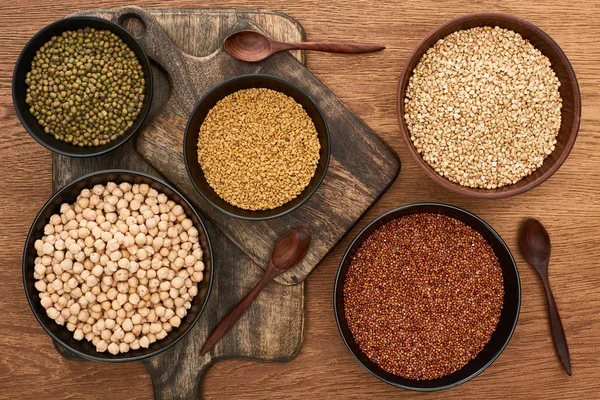 Vista dall'alto di ciotole con fagioli moong, grano saraceno e ceci vicino a cucchiai su taglieri di legno — Foto stock