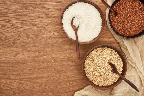 Schalen mit weißem Reis, geröstetem und rohem Buchweizen auf hölzerner Oberfläche mit Leinwand — Stockfoto