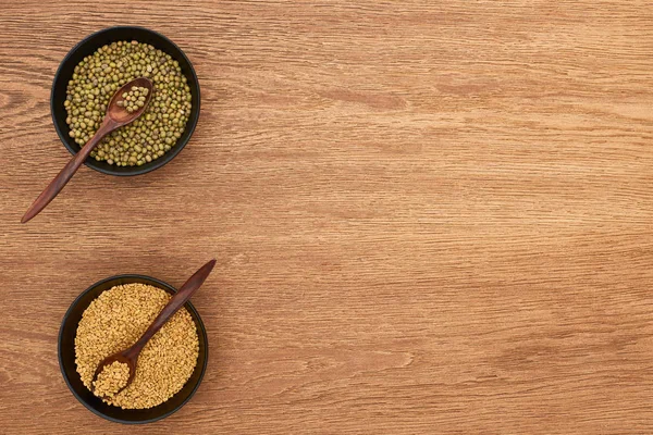 Top view of bowls with moong beans and wheat with spoons on wooden surface — Stock Photo