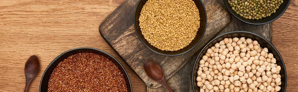Panoramic shot of bowls with buckwheat, beans and chickpea near wooden spoons on cutting boards — Stock Photo