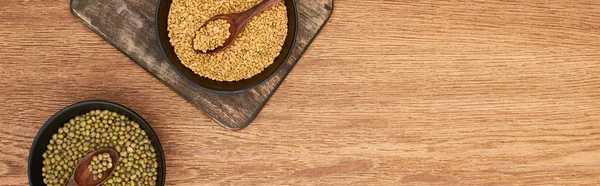 Panoramic shot of black bowls with grains and maash beans with spoons on wooden cutting board — Stock Photo