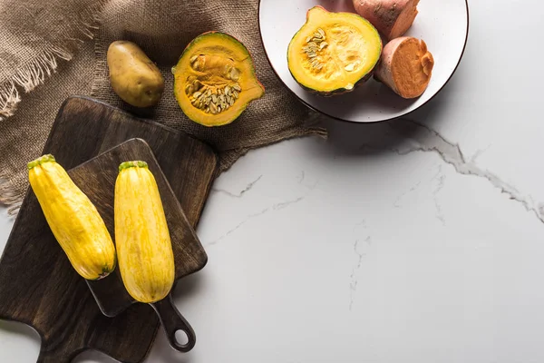 Vista superior de tablas de cortar de madera con calabacín, patata, calabaza y placa con ñame en la superficie de mármol con hessian - foto de stock