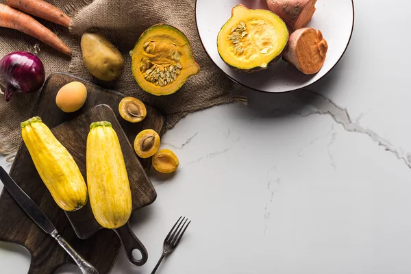 Draufsicht auf Holzschneidebretter und Teller mit Gemüse und Obst in der Nähe von Gabel und Messer auf Marmoroberfläche mit hessischer — Stockfoto