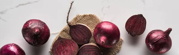 Panoramic shot of red onions and beetroots on marble table with hessian — Stock Photo