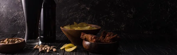 Panoramic shot of glass and bottle of beer near bowls with nuts, crisps and chicken wings on wooden table — Stock Photo