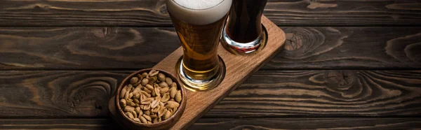 Tiro panorâmico de copos de cerveja escura e leve perto da tigela com amendoins assados na mesa de madeira — Fotografia de Stock