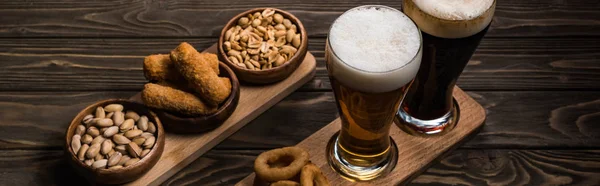 Tiro panorâmico de copos de cerveja escura e leve perto de tigelas com amendoim, pistache, queijo frito e anéis de cebola na mesa de madeira — Fotografia de Stock