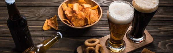 Panoramic shot of glasses of light and bark beer near bottles and snacks on wooden table — Stock Photo