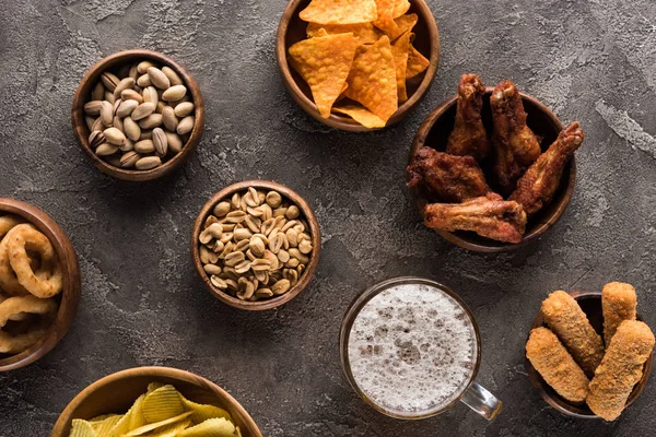 Top view of bowls with nuts, crisps, chicken wings and fried cheese near mug of beer on brown surface — Stock Photo