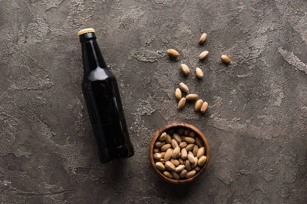 Vista dall'alto di ciotola con pistacchi vicino bottiglia di birra scura sulla superficie marrone — Foto stock
