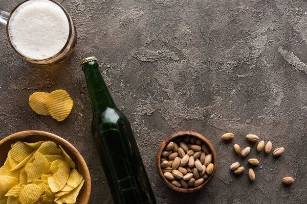 Vista dall'alto di ciotole con pistacchi e patatine vicino bottiglia e tazza di birra sulla superficie marrone — Foto stock