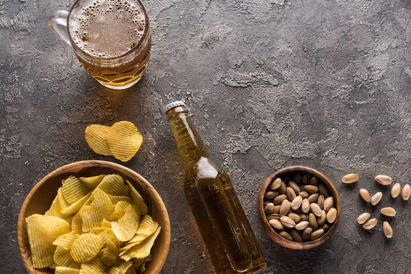 Top view of bowls with pistachios and crisps near bottle and mug of light beer on brown surface — Stock Photo