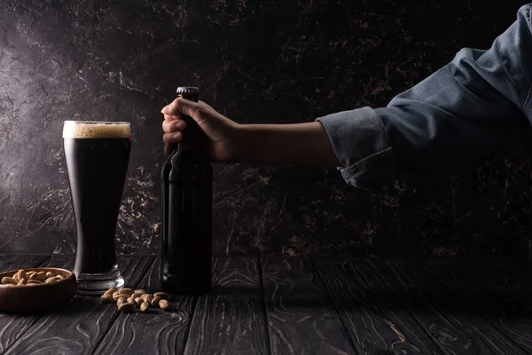 Vue recadrée de l'homme prenant une bouteille près d'un verre de bière et un bol avec des pistaches sur une table en bois — Photo de stock