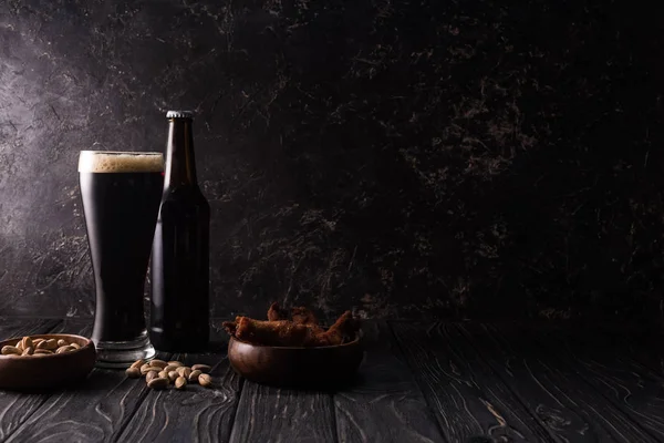 Verre et bouteille de bière noire près des bols avec pistaches et ailes de poulet sur table en bois — Photo de stock