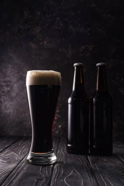 Verre de bière avec mousse près des bouteilles sur table en bois — Photo de stock