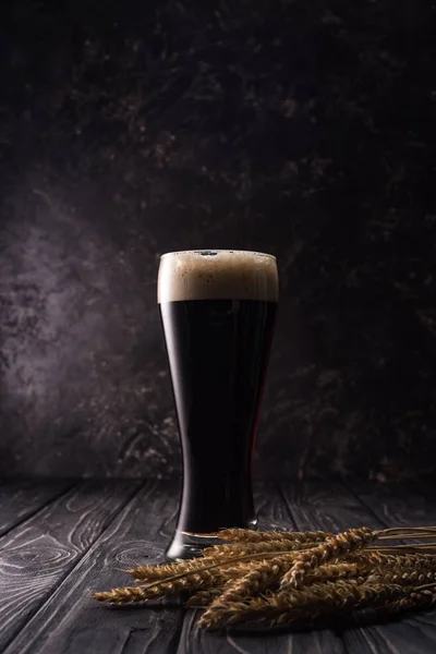 Glass of beer with foam near wheat spikes on wooden table — Stock Photo