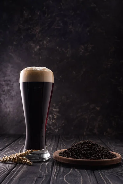 Glass of dark beer near wheat spikes and plate with coffee grains on wooden table — Stock Photo