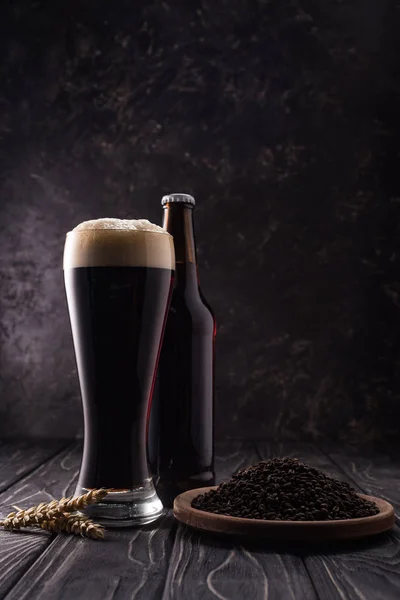 Bouteille et verre de bière près des pointes de blé et assiette avec grains de café sur table en bois — Photo de stock