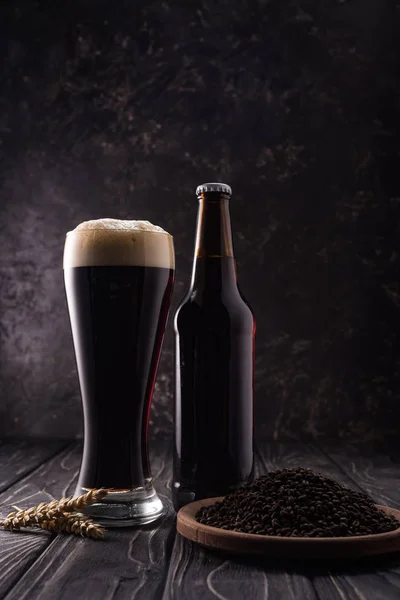 Bouteille et verre de bière noire près des pointes de blé et assiette avec grains de café sur table en bois — Photo de stock