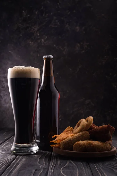Bottiglia e bicchiere di birra vicino al piatto con snack sul tavolo di legno — Foto stock