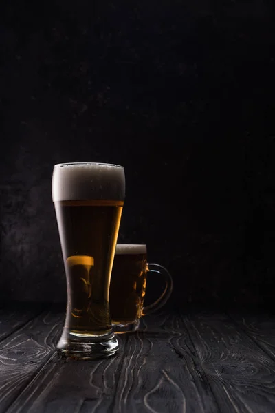 Vidro e caneca de cerveja leve com espuma na mesa de madeira — Fotografia de Stock