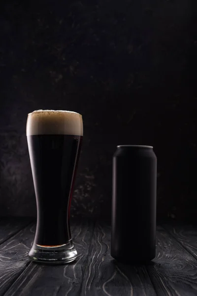 Vetro di birra scura con schiuma vicino lattina nera su tavolo di legno — Foto stock