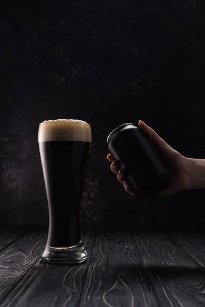 Cropped view of man holding black can near glass of dark beer with foam on wooden table — Stock Photo