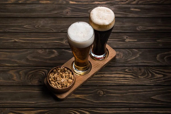 Copos de cerveja escura e leve perto da tigela com amendoins assados na mesa de madeira — Fotografia de Stock