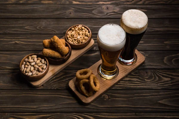 Copos de cerveja escura e leve perto de tigelas com amendoim, pistache, queijo frito e anéis de cebola na mesa de madeira — Fotografia de Stock